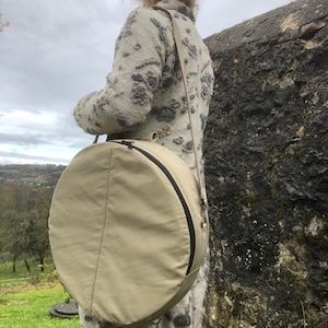 May include: A beige round canvas bag with a zipper and a black strap. The bag is being carried by a person wearing a floral sweater.
