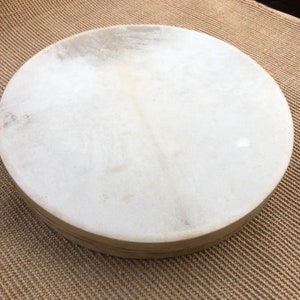 Pode incluir: Um tambor branco redondo com uma cabeça de couro natural. O tambor está sentado em um tapete tecido.