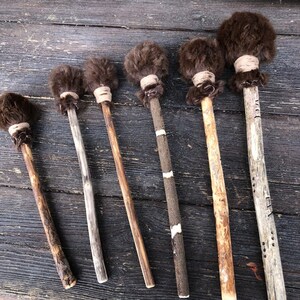 Op de afbeelding: Zes houten drumsticks met bruin bont aan de uiteinden. De drumsticks zijn van verschillende lengtes en hebben natuurlijke houtvariaties.