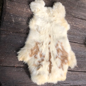 May include: Cream-colored sheepskin rug with brown markings. The rug has a natural shape with rounded edges and a soft, fluffy texture. The sheepskin is laid on a dark wooden surface.