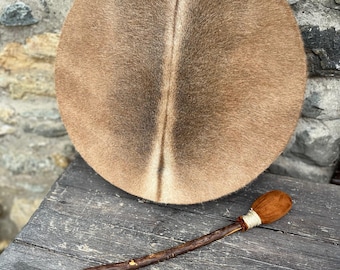 Può includere: Un tamburo di pelle marrone con un bastone di legno per suonare. Il tamburo ha una copertura in pelliccia naturale e una striscia marrone chiaro al centro.