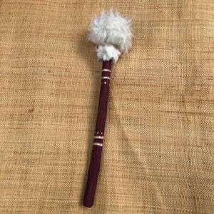 Op de afbeelding: Een houten stok met een wit, pluche pompon aan het uiteinde. De stok is donker bordeauxrood geverfd met witte accenten.