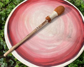 Puede incluir: Un tambor redondo con un marco de color claro y una superficie con círculos concéntricos en tonos rojos y rosas. Un mazo de madera con una cabeza cubierta de tela marrón y un cordón de color claro descansa en diagonal sobre el tambor.