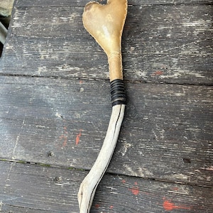 Pode incluir: Objeto artesanal em forma de coração com uma cabeça de couro castanho-claro, uma pega enrolada e uma base de madeira curva. A pega é enrolada com material preto. A base de madeira é de cor clara.