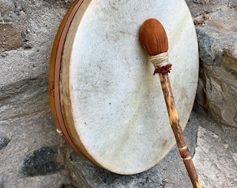 May include: A round, hand-held drum with a light-colored, stretched skin surface. The drum has a wooden frame with a brown and orange trim. A drumstick with a brown, rounded head rests against the drum. The drum and drumstick are set against a stone background.