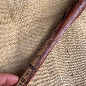 Puede incluir: Una herramienta de madera hecha a mano con un acabado marrón oscuro y diseños grabados intrincados. La herramienta presenta una cabeza redondeada y un mango largo y delgado. El mango tiene una serie de pequeños grabados circulares y otros patrones geométricos.