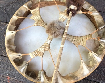 May include: A circular drum with a natural hide head and a wooden frame. The drum is tan and cream, with a brown fur-covered beater. The drum has a rustic, organic aesthetic, with a traditional design.