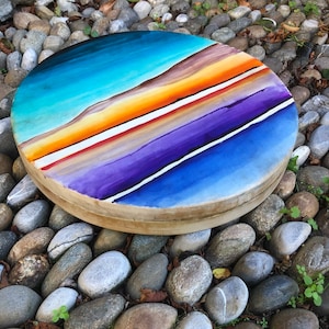 May include: A round wooden frame with a painted design of colorful stripes in shades of blue, purple, orange, and red. The frame is surrounded by small gray stones.