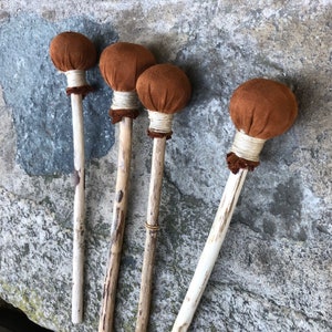 Pode incluir: Quatro baquetas de percussão com cabeças de camurça castanha e cabos de madeira natural. As cabeças são presas com barbante branco e uma faixa de material castanho. As baquetas estão dispostas sobre uma superfície de pedra cinzenta.