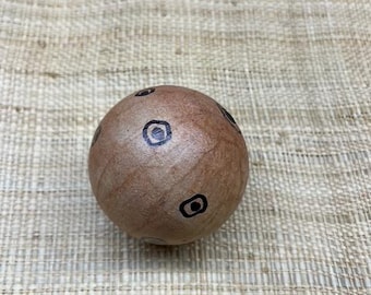 May include: A wooden sphere with a light brown finish and several black, eye-shaped designs. The sphere is resting on a textured, beige surface. The sphere is a decorative object.