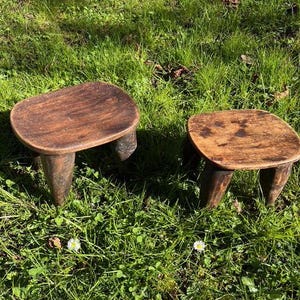 May include: Two small, rustic wooden stools with rounded tops and three sturdy legs each. The stools are a warm brown colour and appear to be handmade. They are set on a bed of green grass.