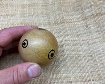 May include: A small, round wooden object with a light brown finish. It features two black, circular eye designs and a small, dark hole on top. The object is held in a hand against a textured, beige background.