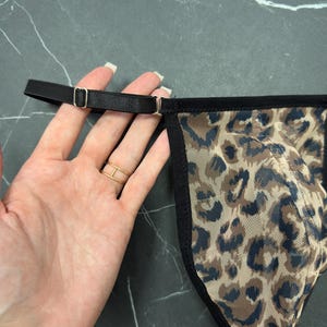 Può includere: Un perizoma con fantasia leopardata nei toni del marrone e del blu. I bordi sono rifiniti con tessuto nero e le cinghie sono nere con finiture metalliche color oro.