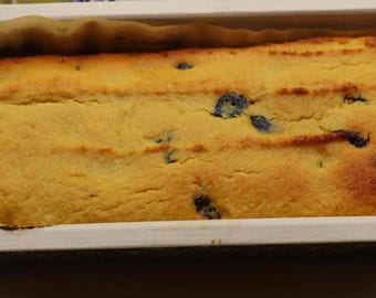 May include: A rectangular loaf of golden-brown baked cake with visible dark blueberries embedded in the surface. The cake is contained within a white paper baking mold lined with parchment paper, showing a textured, porous crumb structure.