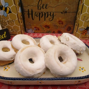 May include: Five white glazed donuts with a hole in the center, arranged on a white oval platter with blue and yellow floral designs. The platter is on a red and white checkered tablecloth.