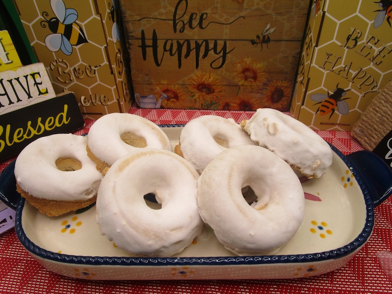 May include: Five white glazed donuts with a hole in the center, arranged on a white oval platter with blue and yellow floral designs. The platter is on a red and white checkered tablecloth.