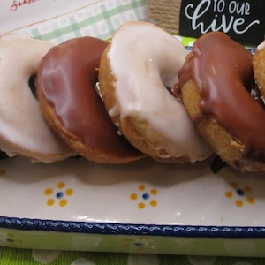 Puede incluir: Una fuente rectangular de cer&aacute;mica con borde azul y detalles florales amarillos contiene seis rosquillas en forma de anillo. Tres rosquillas est&aacute;n cubiertas con glaseado de chocolate con leche y tres con un glaseado blanco opaco.