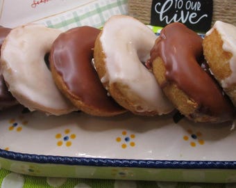 May include: A rectangular ceramic serving dish with a blue rim and yellow floral accents holds six ring-shaped donuts. Three donuts are coated in smooth milk chocolate icing, and three are covered in a thick, opaque white sugar glaze.