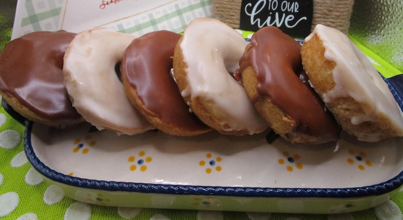 Puede incluir: Una fuente rectangular de cer&aacute;mica con borde azul y detalles florales amarillos contiene seis rosquillas en forma de anillo. Tres rosquillas est&aacute;n cubiertas con glaseado de chocolate con leche y tres con un glaseado blanco opaco.