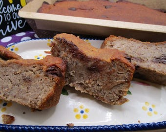 May include: Three slices of banana bread with visible chocolate chips are arranged on an oval white ceramic plate with a blue scalloped edge and yellow flower accents. A loaf of banana bread sits in a paper pan in the background.