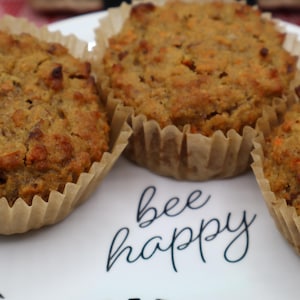 May include: Three golden-brown muffins with visible carrot shreds, each contained in a light tan paper liner. The muffins are arranged on a white plate featuring the black cursive text "bee happy" printed in the center between the baked goods.