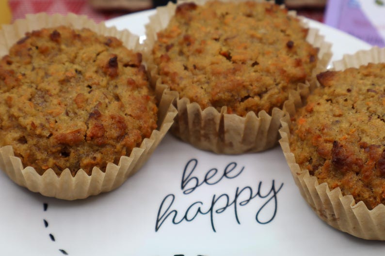 May include: Three golden-brown muffins with visible carrot shreds, each contained in a light tan paper liner. The muffins are arranged on a white plate featuring the black cursive text "bee happy" printed in the center between the baked goods.