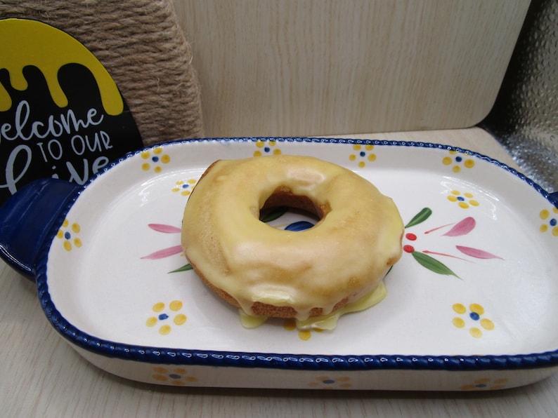 Puede incluir: Un d&oacute;nut glaseado con una cobertura lisa de color tostado claro se sit&uacute;a en el centro de una fuente de cer&aacute;mica ovalada. La fuente tiene una base color crema con motivos florales pintados a mano en azul y amarillo, borde y asas azules.