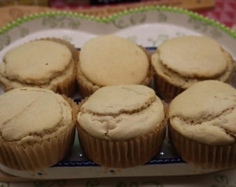 May include: Six light beige muffins with smooth, rounded tops are arranged in two rows of three. Each muffin is contained within a pleated brown paper liner. The muffins are presented on a white serving dish with a decorative green and blue rim.