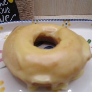 Puede incluir: Un d&oacute;nut redondo con un glaseado suave de color amarillo p&aacute;lido centrado en un plato de cer&aacute;mica blanca que presenta un patr&oacute;n pintado de peque&ntilde;as flores azules y amarillas con hojas verdes. El plato tiene un borde festoneado de color azul.