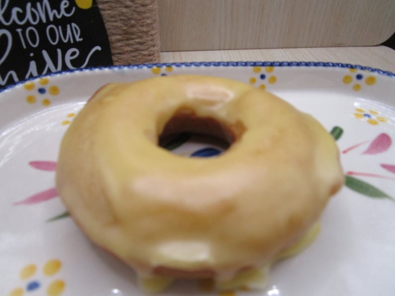 Puede incluir: Un d&oacute;nut redondo con un glaseado suave de color amarillo p&aacute;lido centrado en un plato de cer&aacute;mica blanca que presenta un patr&oacute;n pintado de peque&ntilde;as flores azules y amarillas con hojas verdes. El plato tiene un borde festoneado de color azul.