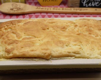 May include: A rectangular loaf of golden-brown baked bread sits inside a light-colored paper baking mold. The top surface of the bread has a textured, uneven crust with a light tan color, while the sides of the mold are smooth and beige.