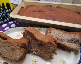 May include: A rectangular loaf of baked bread with visible chocolate chips sits in a paper mold. Three thick slices of the same bread are arranged on an oval ceramic plate with a blue rim and small yellow and blue floral patterns on the surface.