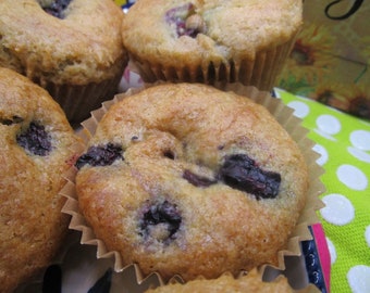 Op de afbeelding: Drie goudbruin gebakken muffins met zichtbare donkerpaarse bosbessen in de bovenkant. Elke muffin zit in een geplooid beige papieren vormpje. De textuur van de bovenkant van de muffins ziet er licht poreus en goudbruin gebakken uit.