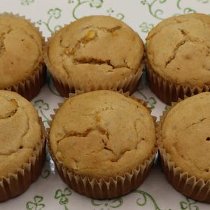 May include: Six golden-brown muffins arranged in two rows of three on a white heart-shaped serving platter. The platter features a green decorative pattern of small clover-like shapes and swirling vines along its scalloped, green-trimmed edge.