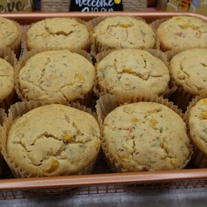 May include: A copper-colored wire basket filled with a dozen golden-brown muffins. The muffins are in paper liners and have a slightly cracked surface, with visible flecks of corn and herbs. The basket is on a white surface.