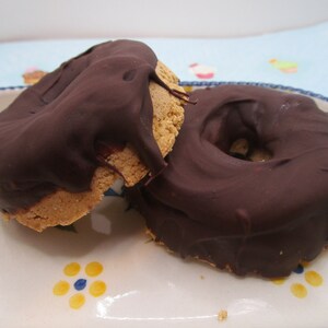 May include: Two chocolate-covered donuts on a white plate with a floral design. The donuts are round and have a hole in the center. The chocolate is dark brown and smooth.
