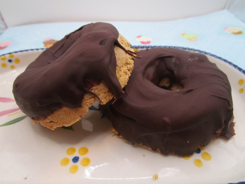 May include: Two chocolate-covered donuts on a white plate with a floral design. The donuts are round and have a hole in the center. The chocolate is dark brown and smooth.