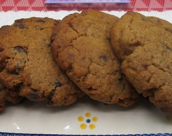 May include: Four large, golden-brown cookies with dark chocolate chips are arranged on a decorative, rectangular serving dish. The dish has a white base with a blue border and a handle. The cookies are baked to a slightly cracked texture.