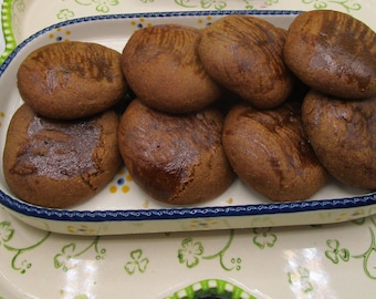 May include: A serving of freshly baked, round, brown cookies arranged in a decorative, oval-shaped ceramic dish. The dish is white with a blue rim and handles, set on a larger tray with a green floral pattern. The cookies have a slightly textured surface.