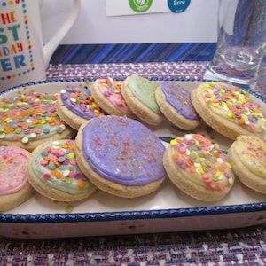 May include: A rectangular serving dish filled with frosted sugar cookies. The cookies have pastel-colored frosting and are decorated with colorful sprinkles. A white mug with the text "HAVE THE BEST BIRTHDAY EVER" is in the background.