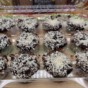 May include: A clear plastic container holding a dozen mini chocolate donuts. The donuts are covered in chocolate frosting and topped with shredded coconut. The donuts are arranged in rows of three.