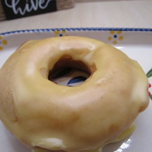 Puede incluir: Un d&oacute;nut redondo de color marr&oacute;n dorado con un glaseado suave de color amarillo p&aacute;lido que cubre la superficie superior. El d&oacute;nut tiene un agujero central y est&aacute; colocado sobre un plato de cer&aacute;mica blanca decorado con flores azules y amarillas.