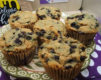 Könnte beinhalten: Eine Ansammlung von fünf gebackenen Blaubeermuffins auf einem dekorativen Teller. Jeder Muffin hat eine goldbraune, strukturierte Oberfläche mit sichtbaren dunkelblauen Beeren, die im Teig eingebettet sind, und steckt in einer braunen Papierform.