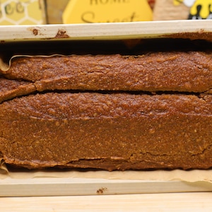May include: A rectangular loaf of dark brown baked bread sits inside a light-colored wooden baking mold. The bread has a textured, cracked surface and is lined with brown parchment paper along the bottom and sides of the wooden container.