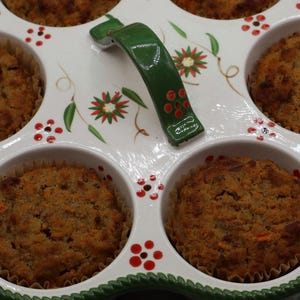 May include: A white ceramic muffin pan with six circular wells, each containing a baked brown muffin. The pan features a green handle and a decorative pattern of small red flowers and green leaves. The rim has a textured green rope-like border.