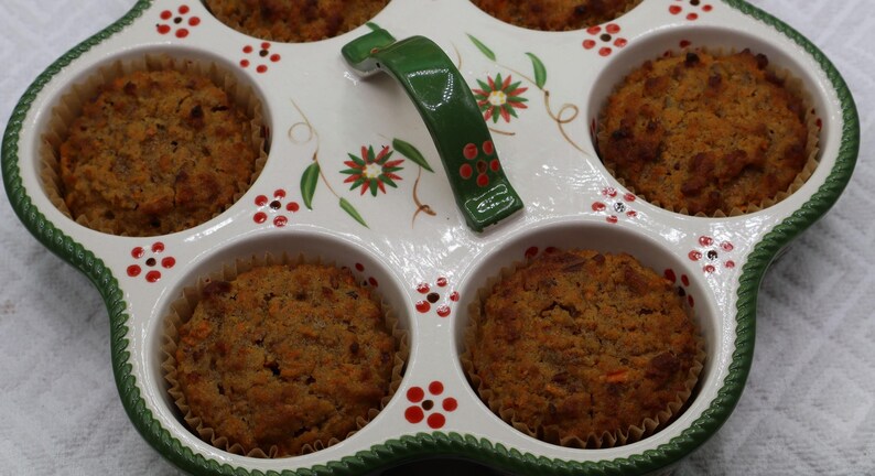 May include: A white ceramic muffin pan with six circular wells, each containing a baked brown muffin. The pan features a green handle and a decorative pattern of small red flowers and green leaves. The rim has a textured green rope-like border.