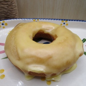 Puede incluir: Un d&oacute;nut redondo de color marr&oacute;n dorado cubierto con un glaseado suave de color amarillo p&aacute;lido que gotea por los lados. El d&oacute;nut descansa sobre un plato de cer&aacute;mica blanca decorado con un patr&oacute;n de peque&ntilde;as flores amarillas con centros azules y hojas verdes.
