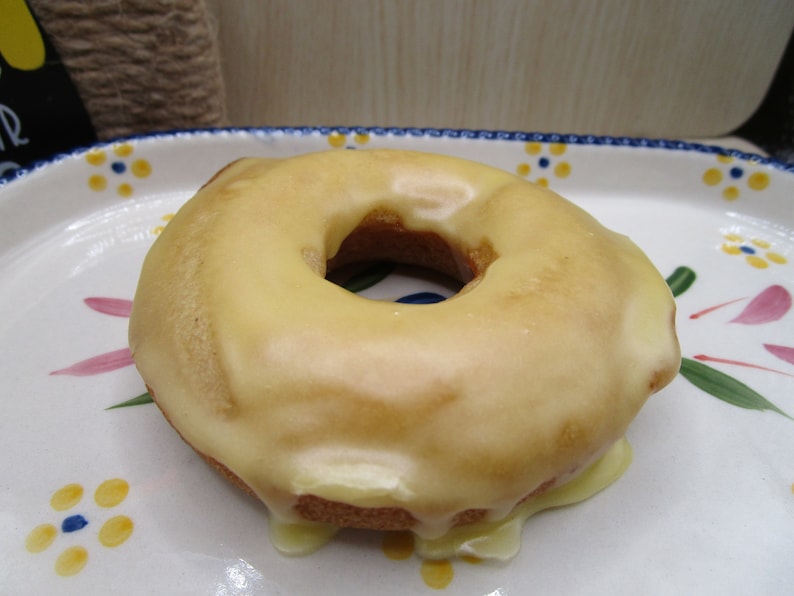 Puede incluir: Un d&oacute;nut redondo de color marr&oacute;n dorado cubierto con un glaseado suave de color amarillo p&aacute;lido que gotea por los lados. El d&oacute;nut descansa sobre un plato de cer&aacute;mica blanca decorado con un patr&oacute;n de peque&ntilde;as flores amarillas con centros azules y hojas verdes.
