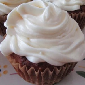 May include: A single cupcake featuring a golden-brown baked base held in a pleated brown paper liner, topped with a generous, thick swirl of smooth, opaque white frosting that covers the entire top surface of the cake in a decorative pattern.
