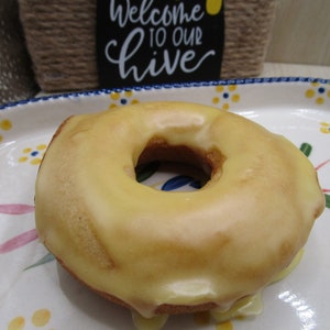 Puede incluir: Un d&oacute;nut redondo de color marr&oacute;n dorado con un glaseado suave de color amarillo p&aacute;lido, sobre un plato ovalado de cer&aacute;mica blanca decorado con peque&ntilde;os motivos florales azules y amarillos y p&eacute;talos rosas. Se ve un cartel con Welcome to our hive.